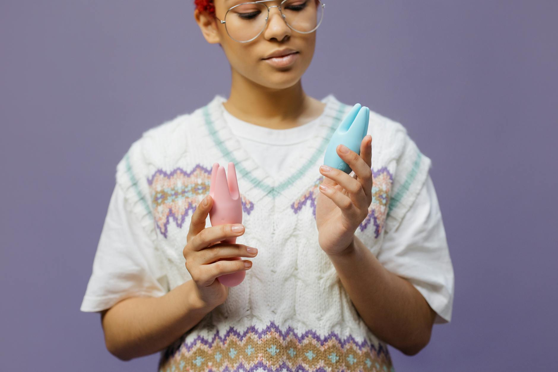 Woman with eyeglasses holding blue and pink silicone vibrators in a contemplative manner while considering intimate connection.